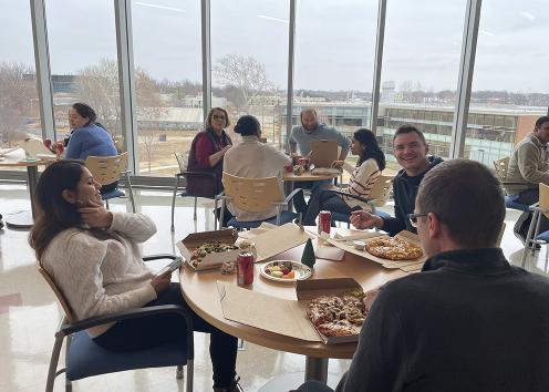  Group eating pizza at tables or 3 or 4 in front of large windows overlooking winter landscape from 3rd floor of MRB interaction area.